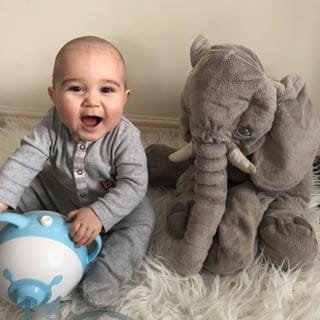 Ein heiterer Babyjunge sitzt auf einem Bett neben einem Plüschelefanten und hält den bärenförmigen Nosiboo Pro elektrischen Baby Nasensauger an den Ohren fest
