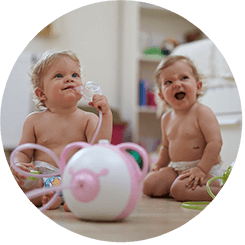 Zwei glückliche Babys sitzen auf dem Fußboden mit dem Nosiboo Pro elektrischen Baby Nasensauger