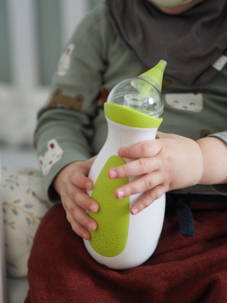 Ein süßer kleiner Junge, der auf dem Bett sitzt und den tragbaren Baby Nasensauger Nosiboo Go in den Händen hält