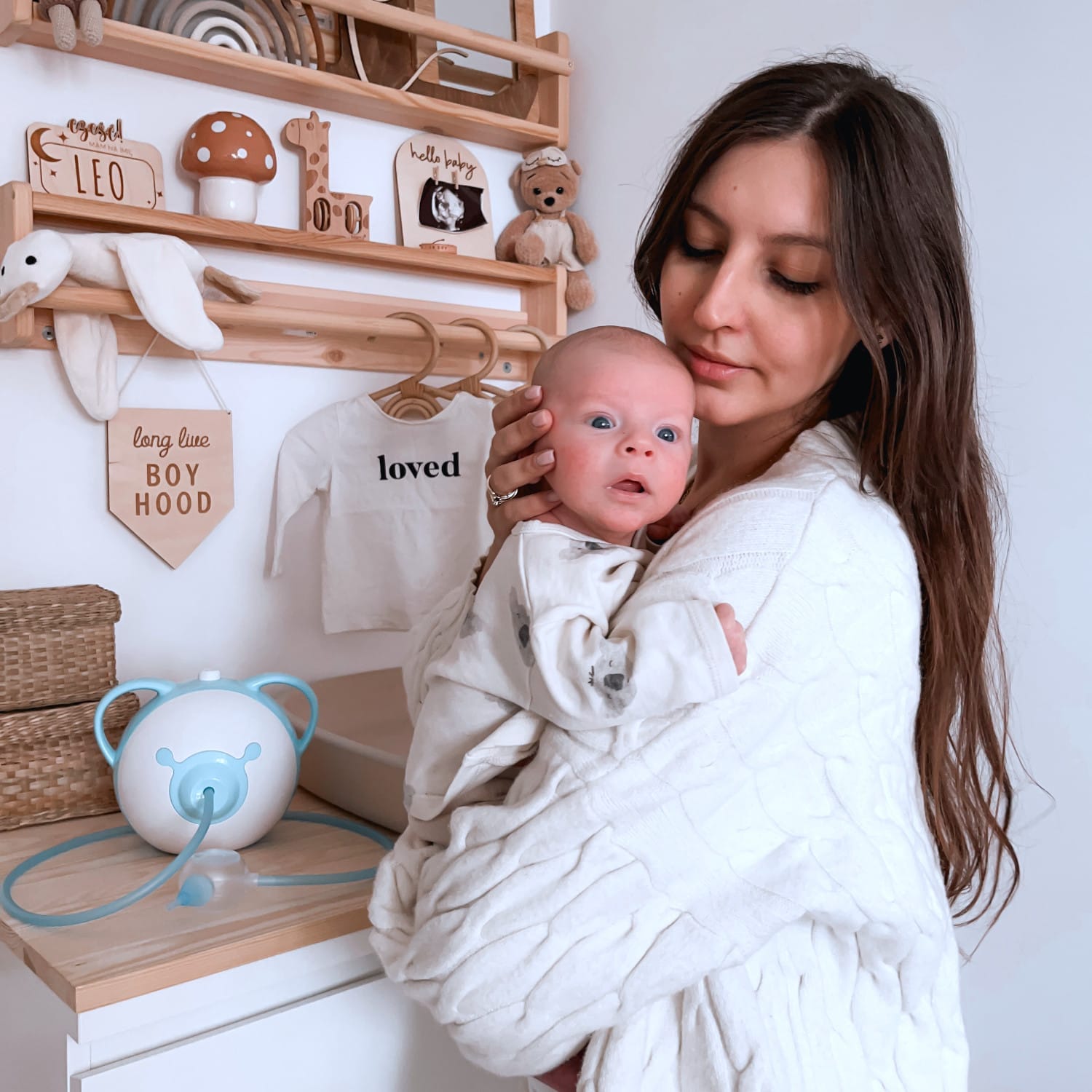 Eine hübsche Mutter hält ihr süßes neugeborenes Baby in ihren Armen, während sie in einem Kinderzimmer neben einem Nosiboo Pro elektrischen Nasensauger steht.